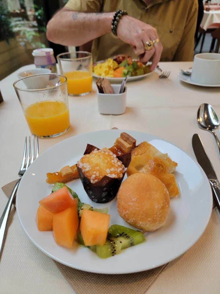 Two breakfast plates with mini-muffins, focaccia, canteloupe and kiwi, two glasses of orange juice, and coffee