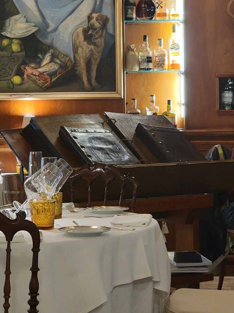 Two large leather-bound books sit atop another extremely large open ring binder with thick leather pages. A table set for dinner with a white tablecloth in the foreground.