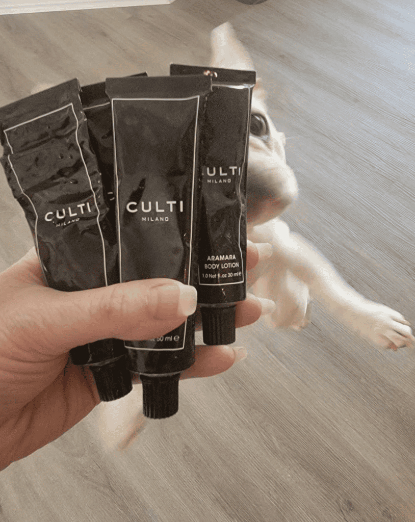 A woman's hand holds black tubes of Culti bath and bodycare products while a white french bulldog leaps up to see them closer