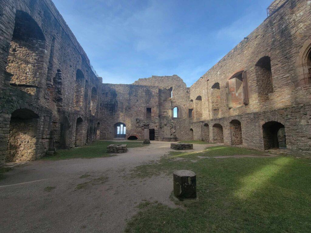 A large open area partially covered in grass with small stone structures, surrounded by high stone walls with arches and windows