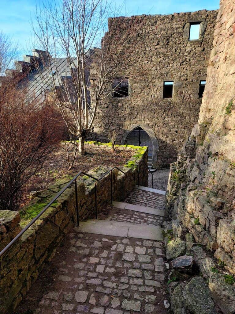 A stone path leads to shallow stairs. A stone wall on the right is in a partial state of decay. In the back a stone wall with windows and an arched doorway