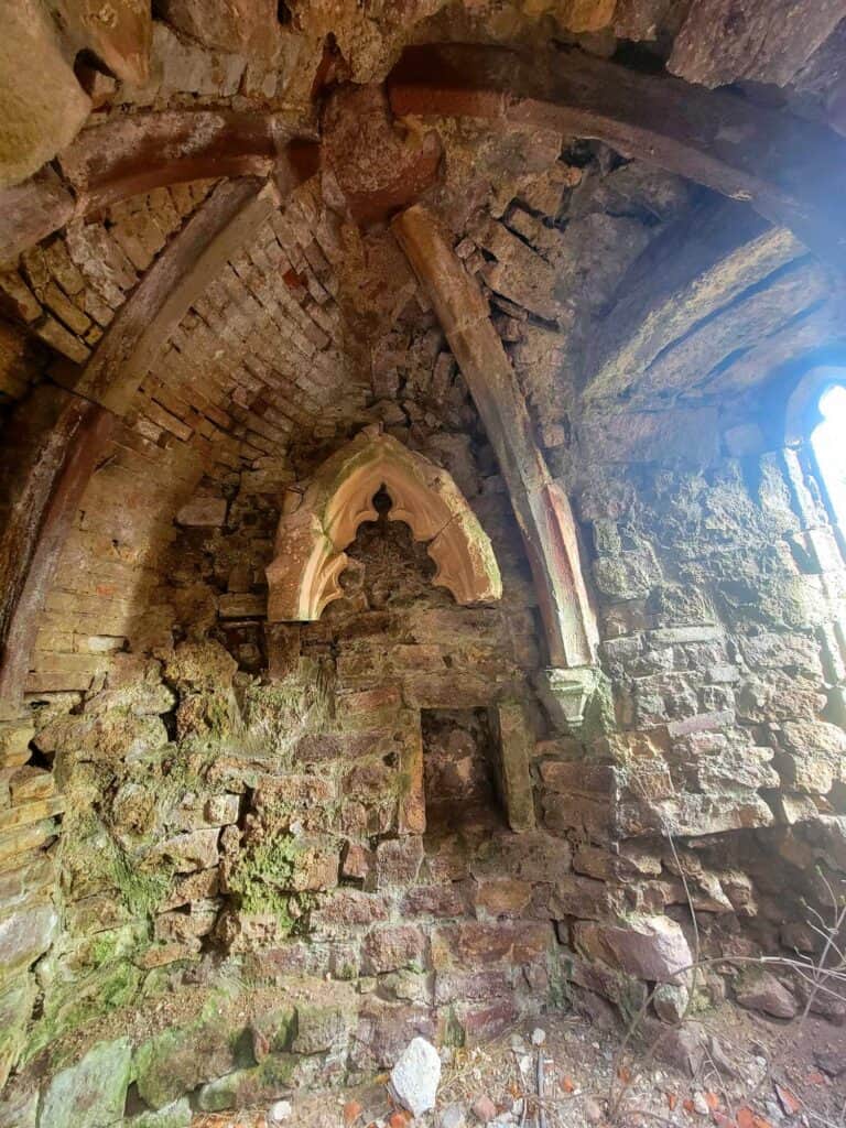 A small stone room with a braced dome ceiling, moss covered rocks, and an ornamental window casing.