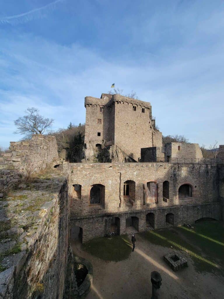 A view down into an open grassy area with a stone well, and a wall with arches. Above is a rectangular upper castle with ramparts