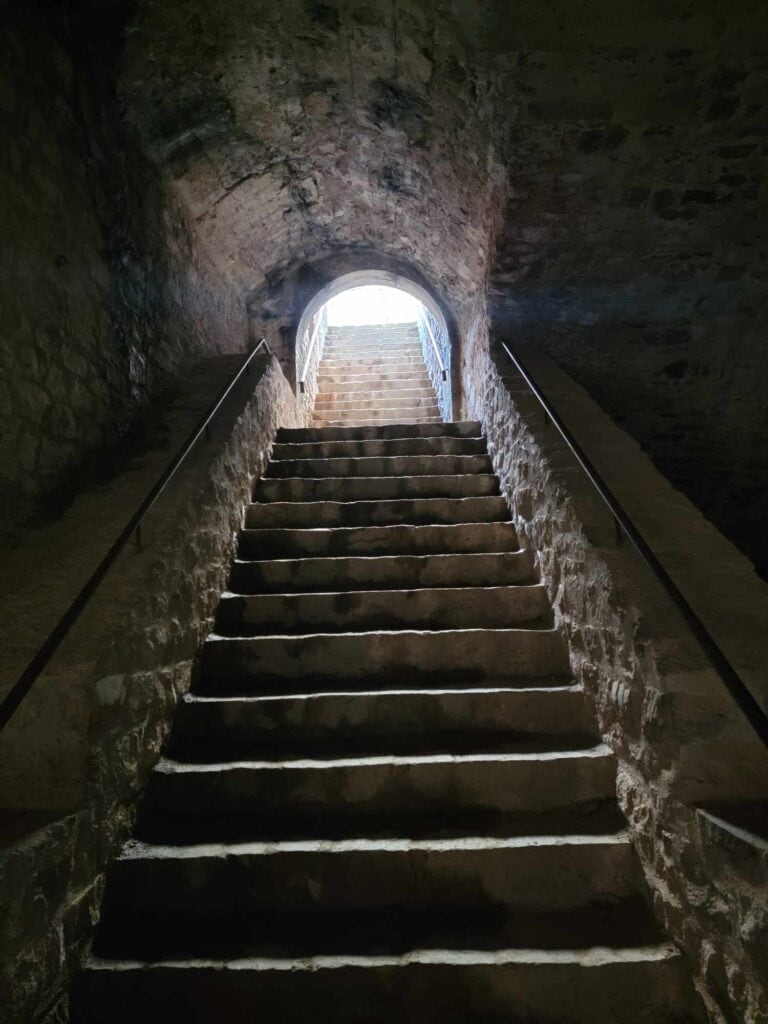 A long set of stone stairs with metal railings inside a rounded tunnel with sunlight at the top