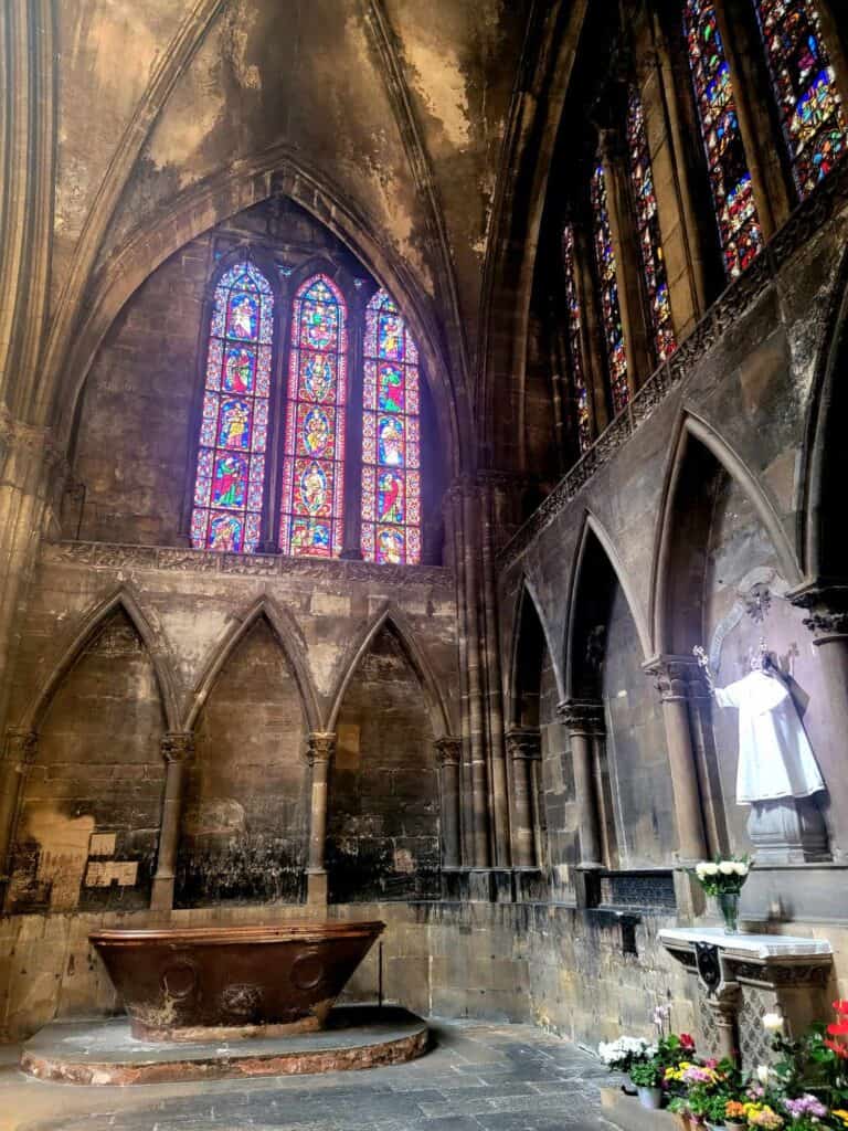 The corner of a stone cathedral, walls covered in soot. A medieval stained glass triptych over a wall with high pointed arches. A bronze tup, sloped on one side and a lion head to the left.