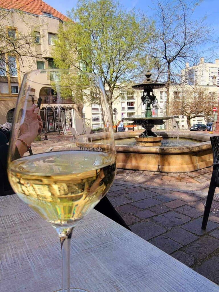 A glass of white wine sits on a table with a fountain in the background