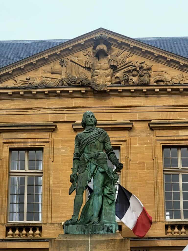 A green bronze statue of a man in a cloak with a stone building behind flying the French flag