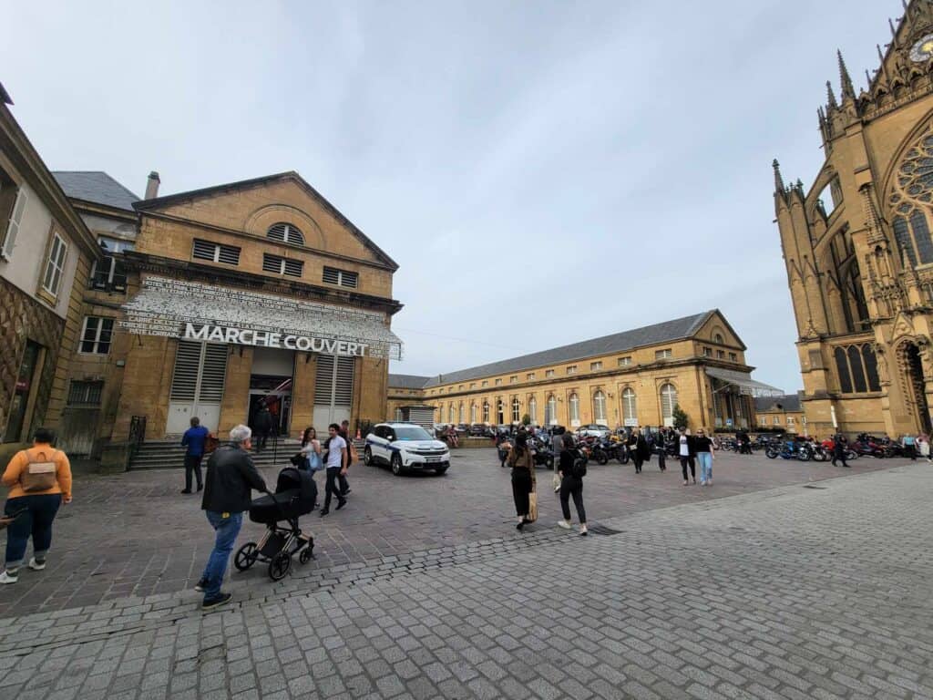 A cobblestone square with a U-shaped building of yellow stone and a sign that reads Marché Couvert. People walking past, and the cathedral to the right