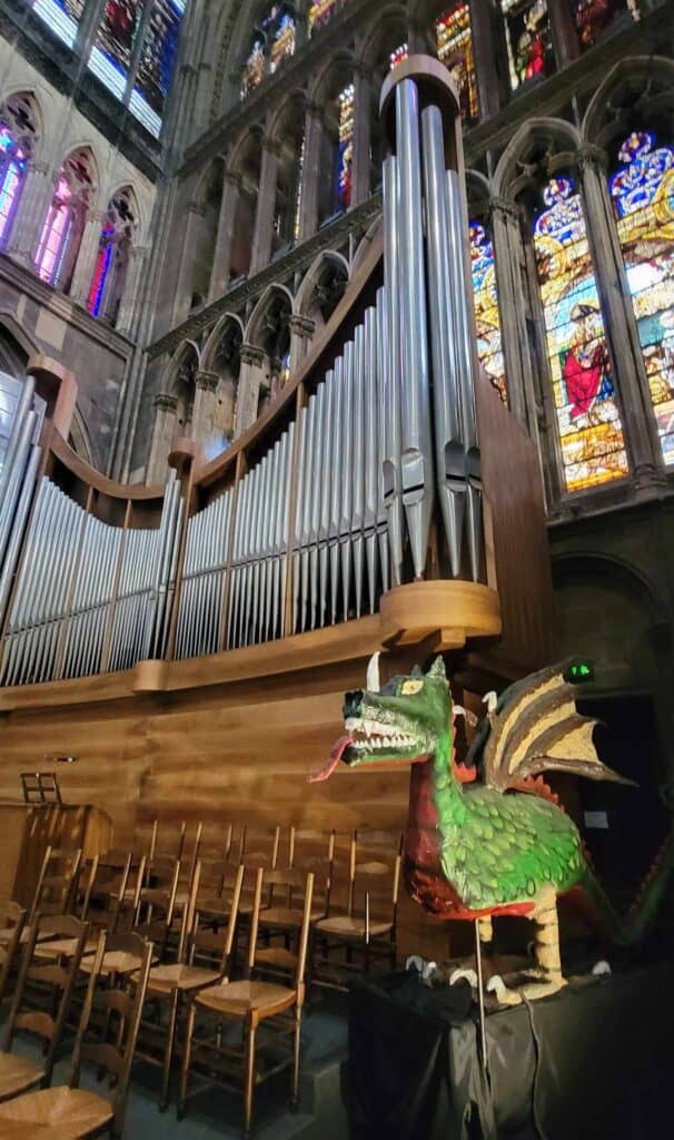 A green paper maché dragon with yellow legs and a horn on its nose stands by wooden chairs with a large pipe organ behind