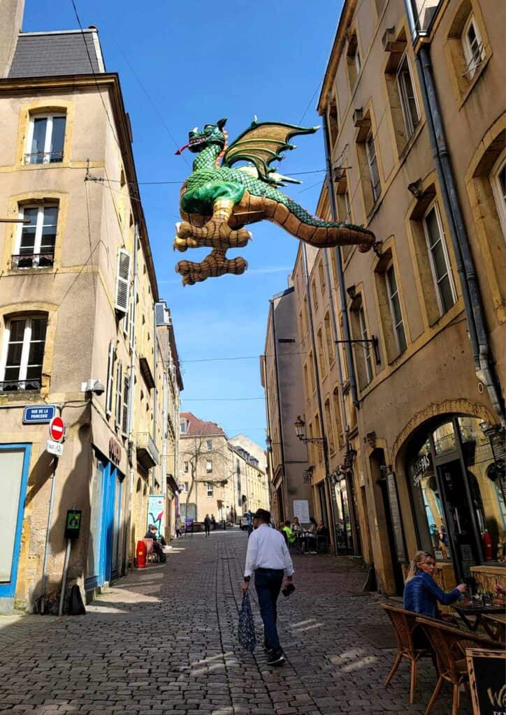 A man walks down a cobblestone street lined with buildings and a large winged dragon overhead