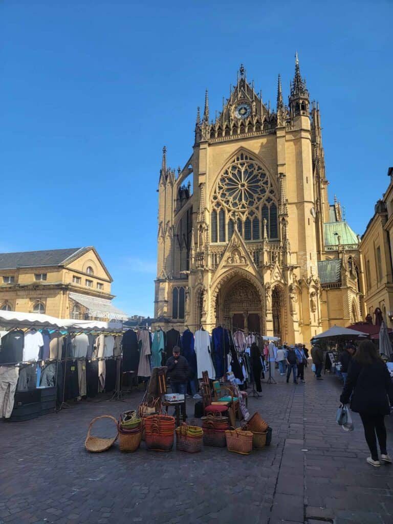 A tall cathedral front with a rose window and a large clock on the topmost point. Stalls selling baskets and clothing in the square in front