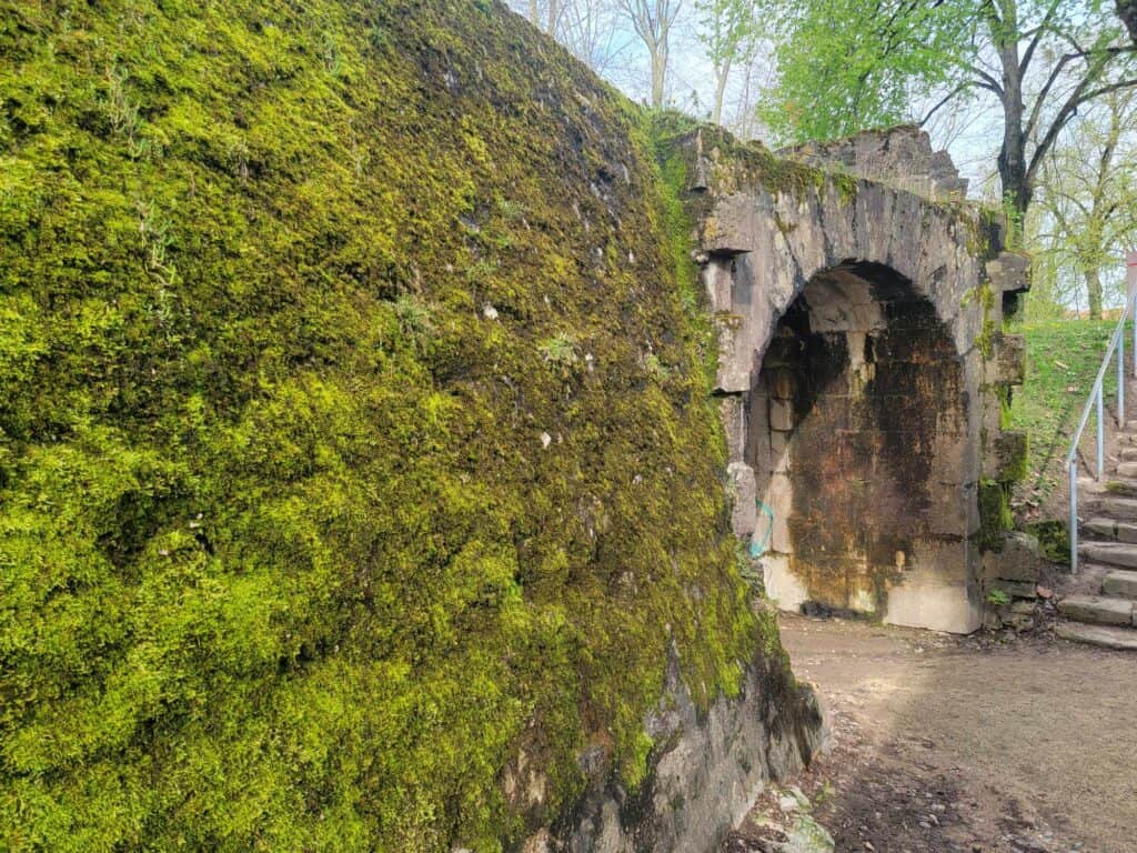 A stone wall covered in moss with a rounded passage way through it and stairs leading up to a path