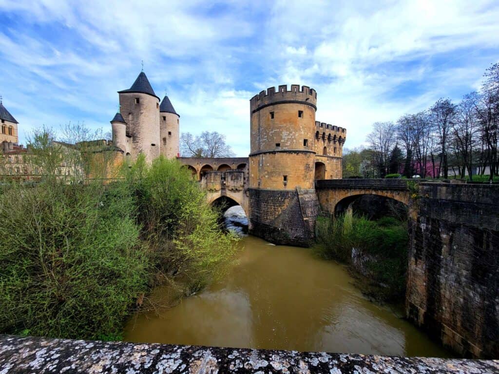 An old stone bridge with arches crossing a narrow brownish colored river, a pale set of three stone towers with black conical slate roofs on the left and a rounded orange stone castle with small windows on the right.