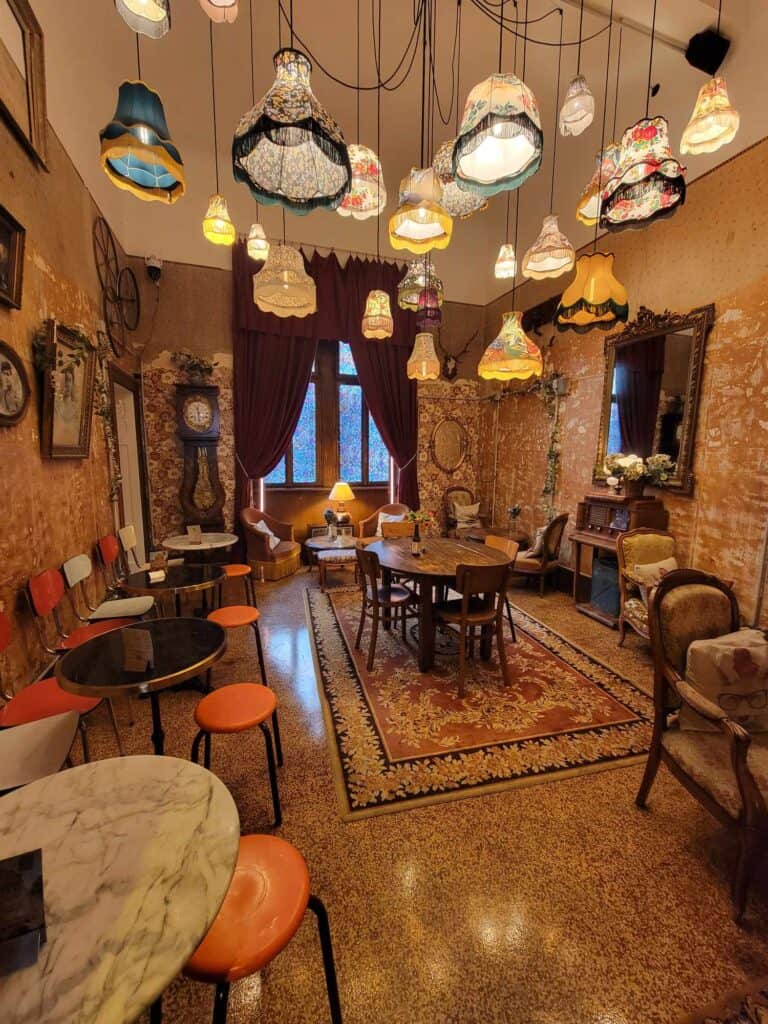 A beautiful richly decorated room with textured warm toned wallpaper and more than 20 light fixtures with different cloth fringed shades hanging from the ceiling Marble café tables with orange stools, and upholstered wooden chairs with a large 19th century style area rug