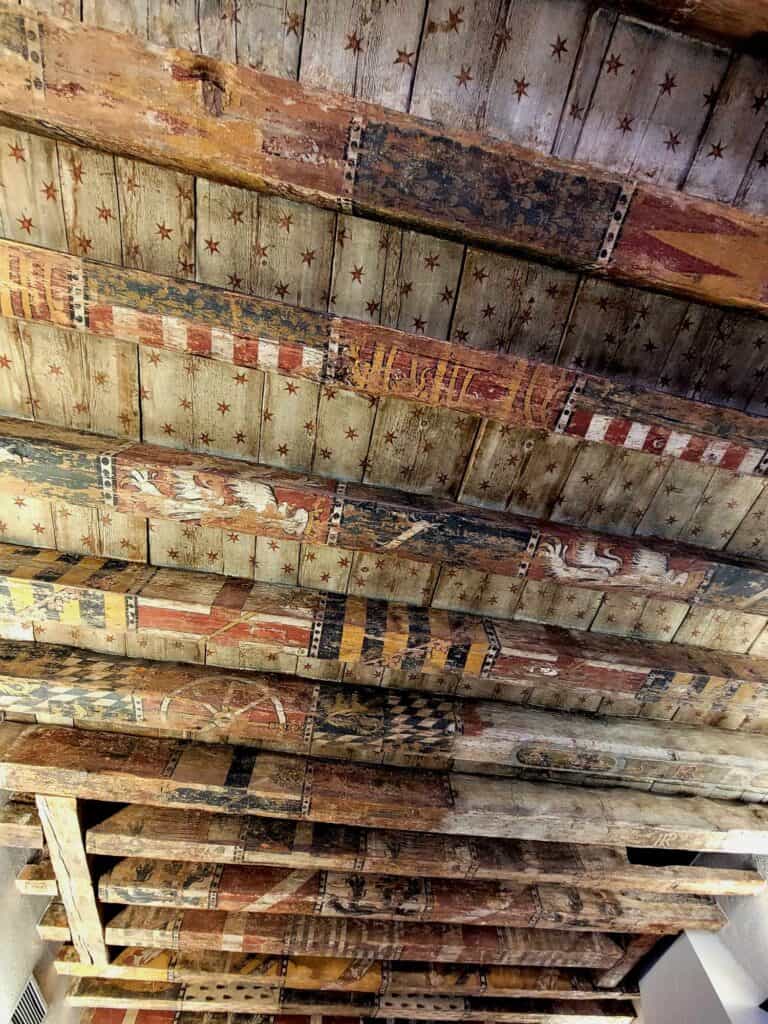 A ceiling with joists painted in blocked designs of stars, stripes, chevrons, wheels and other geometric shapes in red, blue, yellow, black and white