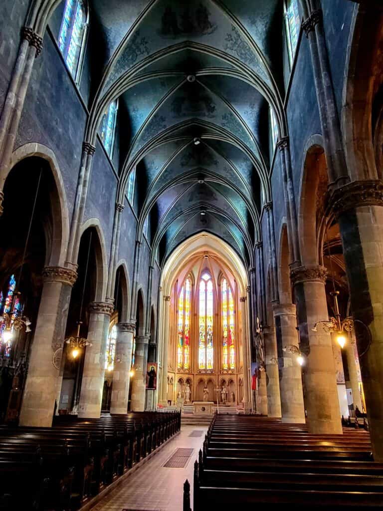 A tall nave with a painted ceiling, rows of pews and an altar surrounded by gold and red stained glass