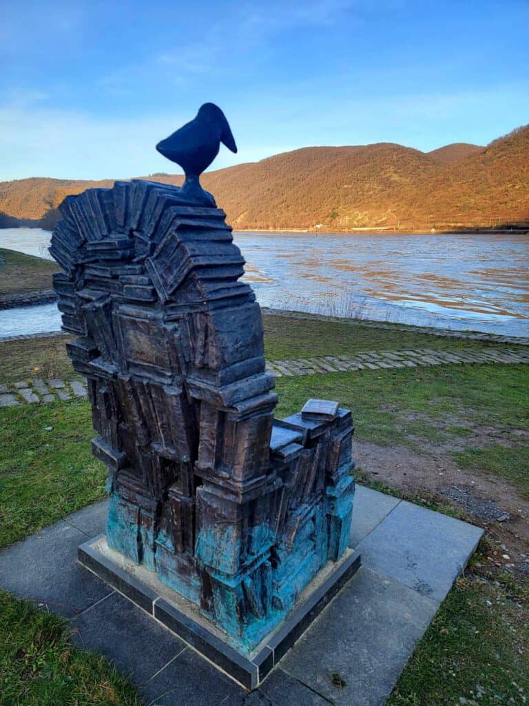 A bronze chair with a high back made to look like it was made from stacks of books. A bronze raven sits on top