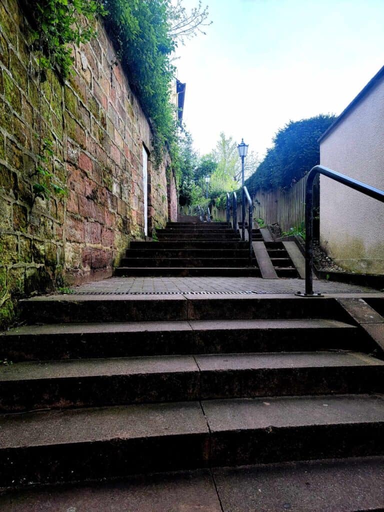 Wide steps leading up with a handrail and a stone wall on the left