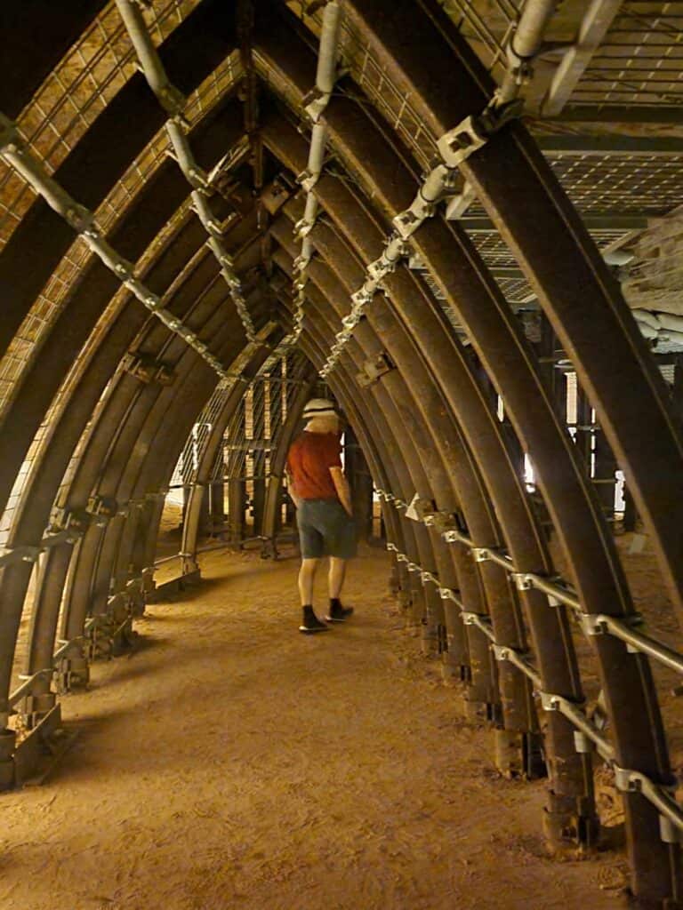 A man wearing a hard hat walks under a series of steel arches on a red sandy path