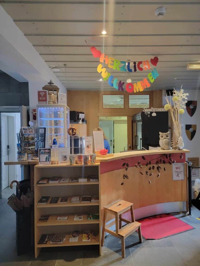 A light wood curved reception desk with a bookcase next to it holding pamphlets. Rainbow colored letters above spell Herzlich Wilkommen with hearts on either side