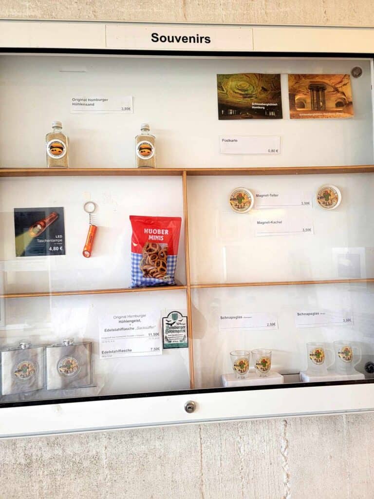 A glass cabinet labeled "souvenirs" holding a sparse configuration of postcards, magnets, flasks, a flashlight and a bag of mini pretzels