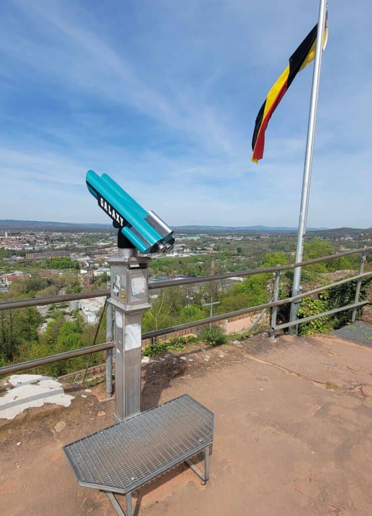 Turquoise binoculars on a metal stand looking over a suburban landscape with a German flag on a flagpole to the right