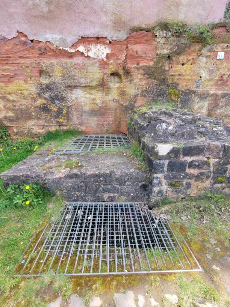 An eroding wall with a large hole in front covered with a metal grate