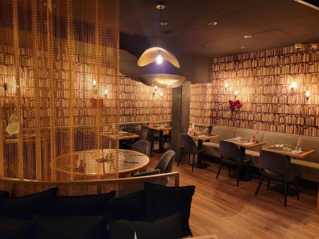 A room with moody lighting and tables with grey bench seating and upholstered chairs and wallpaper that looks like rows of books