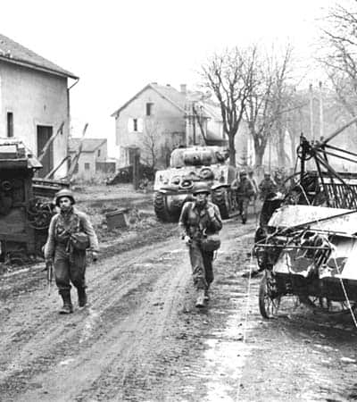 A grainy black and white photo of two US Army GIs walking toward the camera on a muddy road. Other soldiers farther in the back walking past a disabled tank and other machinery.