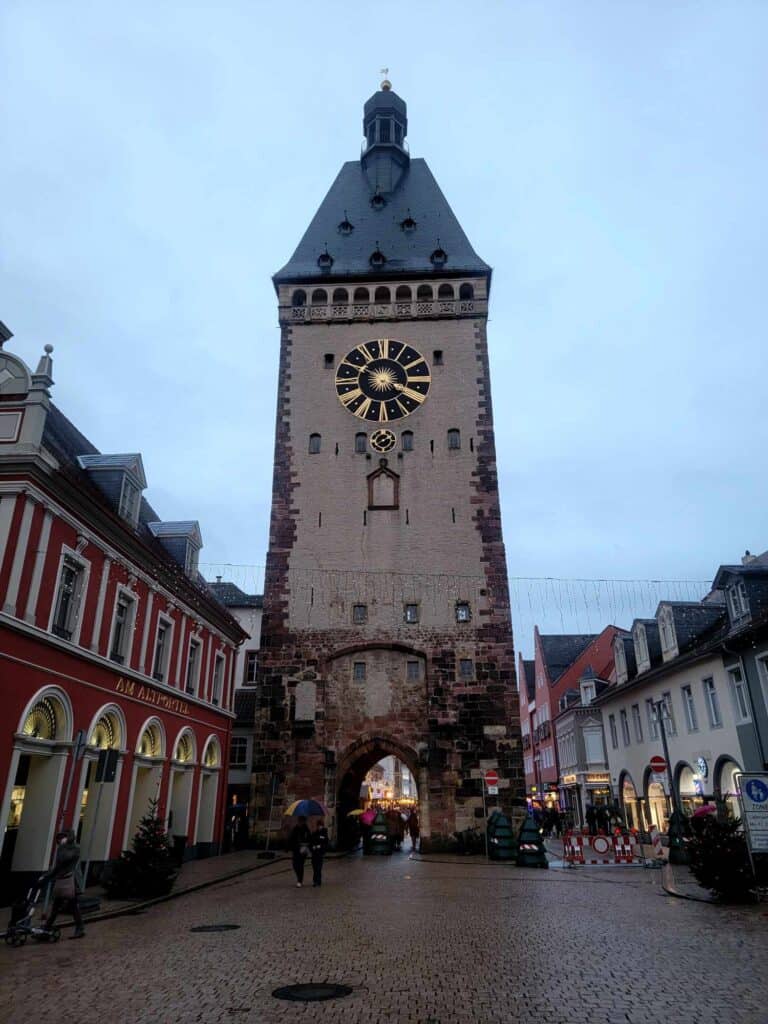 A tall Medieval tower with an arched passage through the center on the street, featuring a clock and pointed slate roof