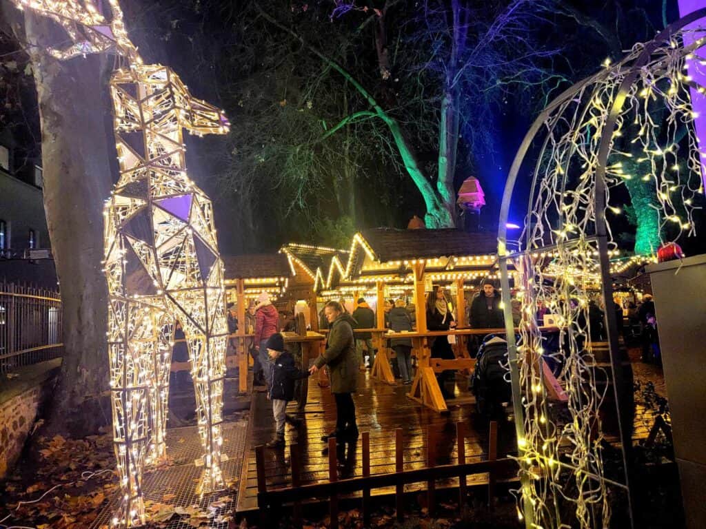 Illuminated wooden shelters with tables and people standing and eating, with large reindeer in tiny white lights on the side