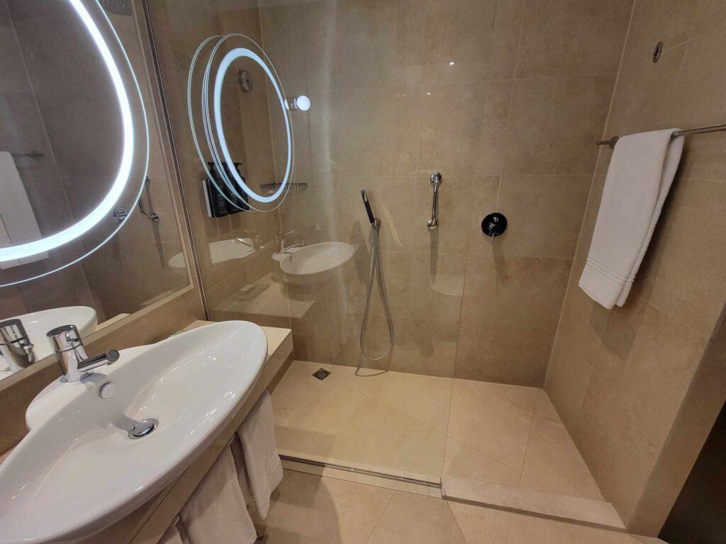 A clean modern bathroom with a circular lighted mirror and a large oval sink in front of a glass-doored shower