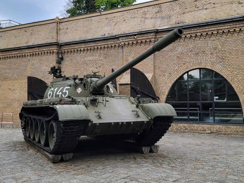 A large olive green tank with 6145 written on the side in white. It sits in front of a yellowish brick building with arched windows