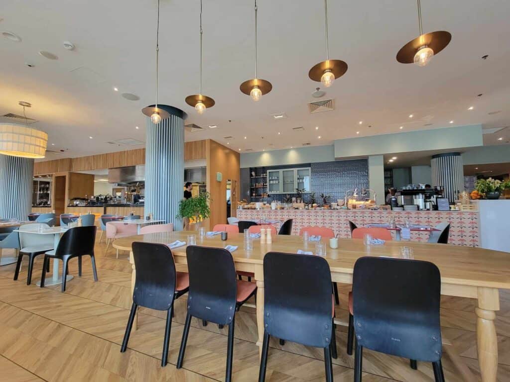 Dark chairs sit pushed in under a long pale wood table set for breakfast with a large buffet in the background