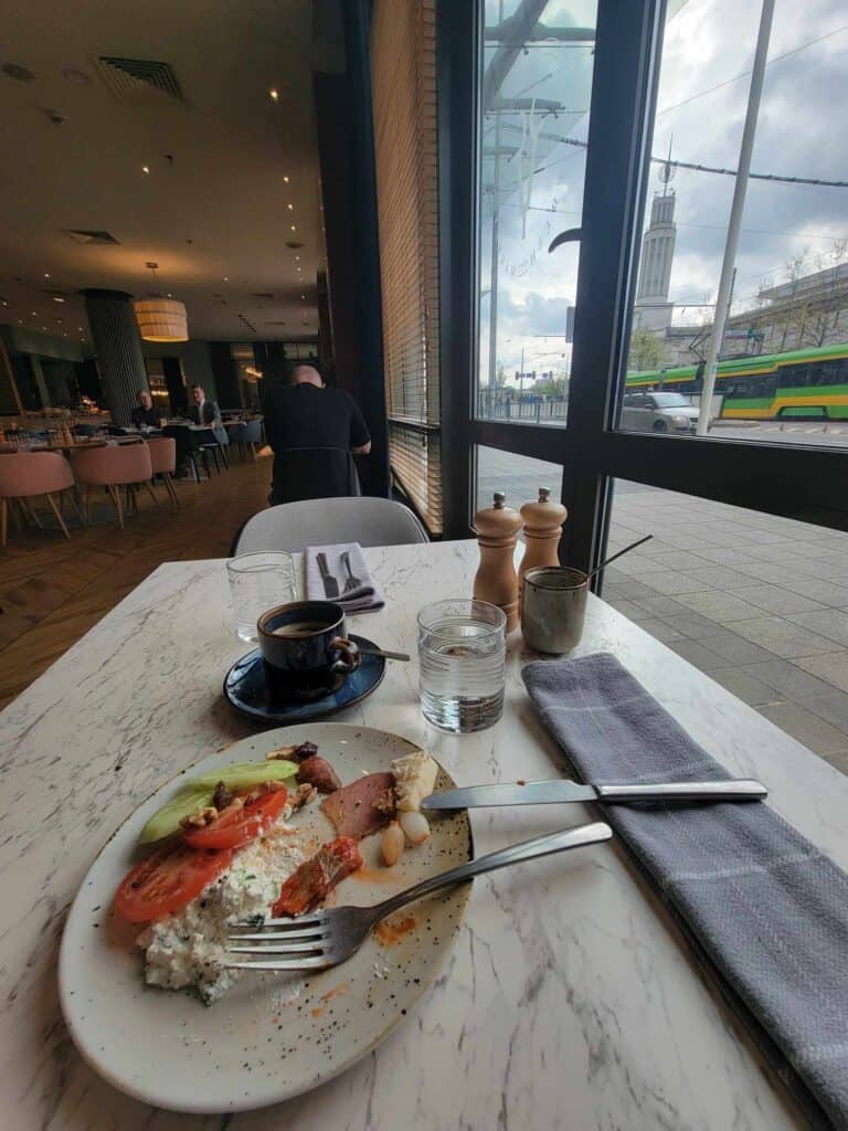 A plate with cucumber, tomato, herring, and cottage cheese sits next to a cup of coffee on a marble table next to a window