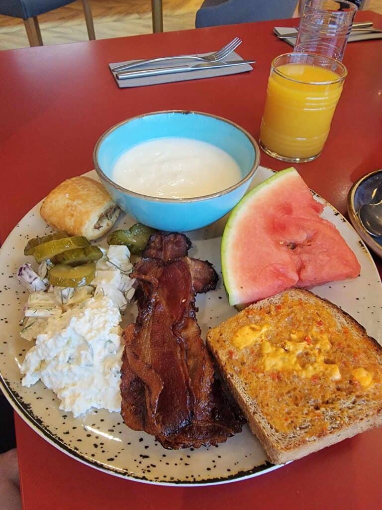 A plate with bacon, cottage cheese, watermelon, a bowl of yogurt,pickles, onion pastry, and toasted wheat bread with tomato butter sits next to a glass of orange juice.