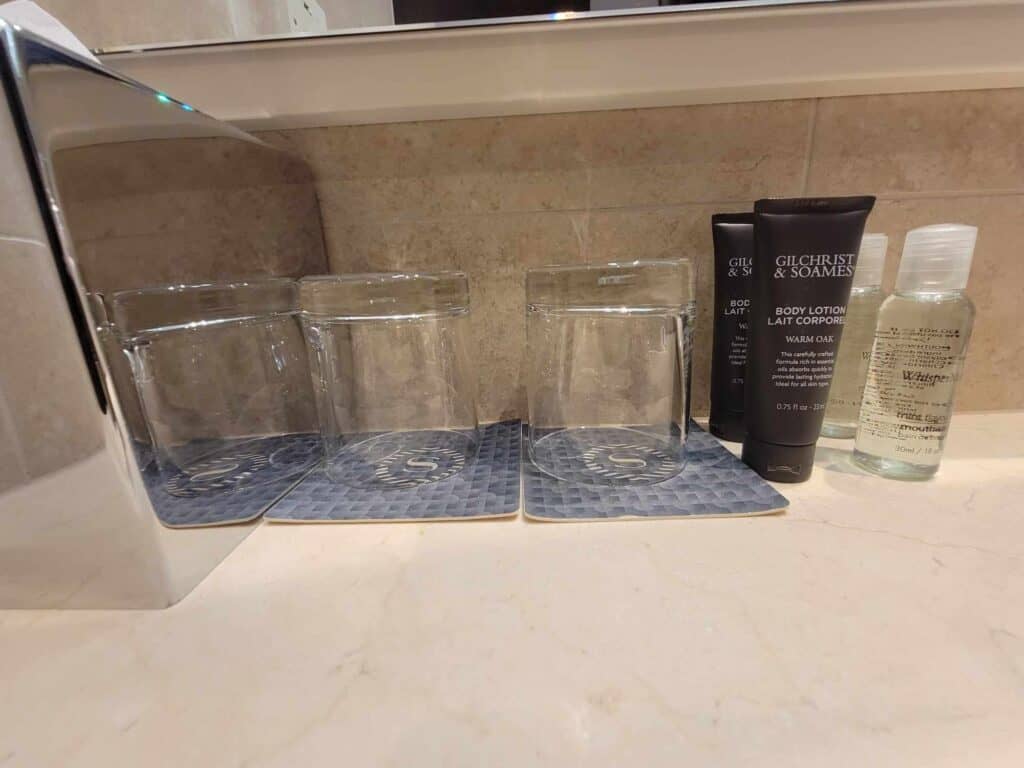 Two glasses sit on a marble countertop next to small black containers of shampoo and lotion