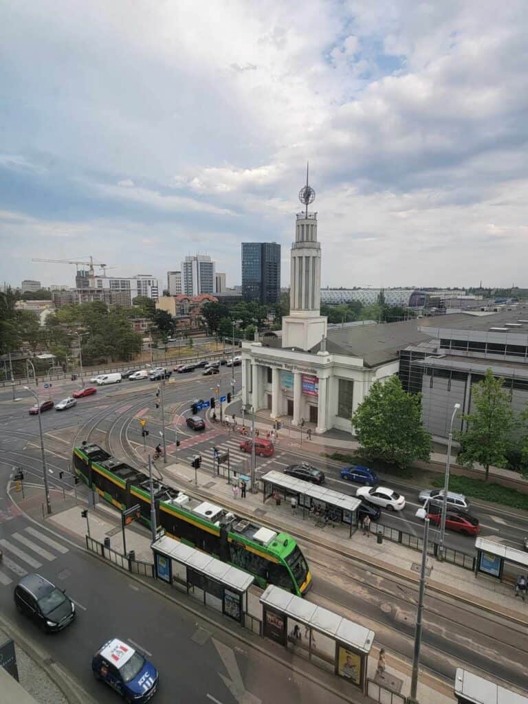 A white building with columns on the front and a tower with a spire on which sits a small metal globe. A green streetcar and traffic pass by