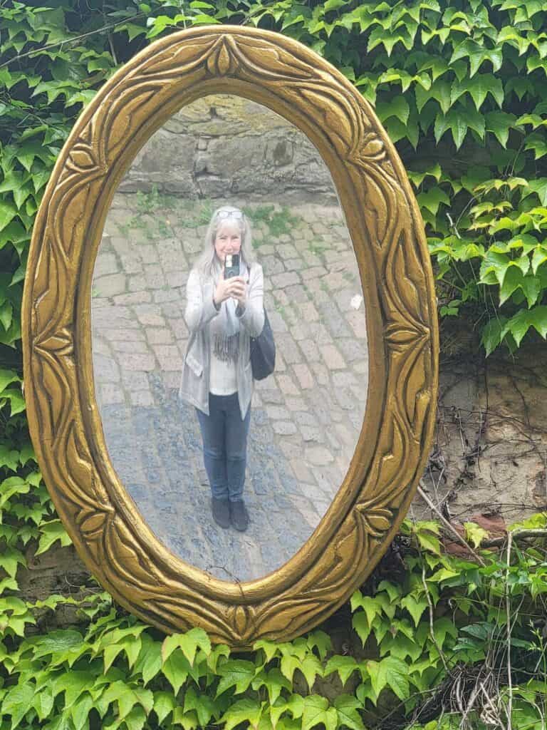 A woman taking a selfie in a large overhead oval mirror on a wall covered with ivy