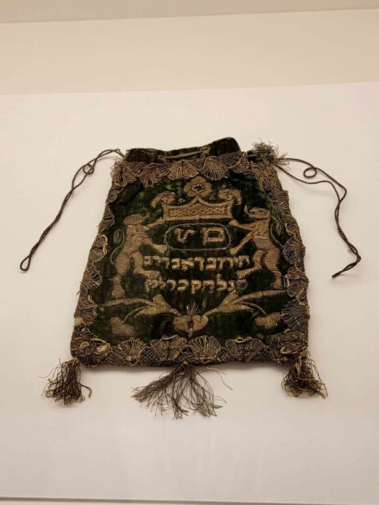 A dark purple velvet pouch with a drawstring and tassles on the corners with embroidered decoration and Hebrew letters