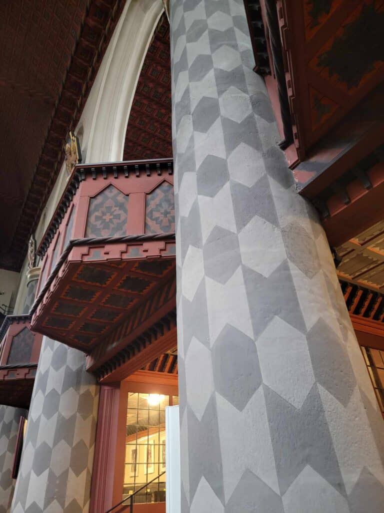 A church interior with geometric designs painted on wide tall stone columns and a red wooden balcony