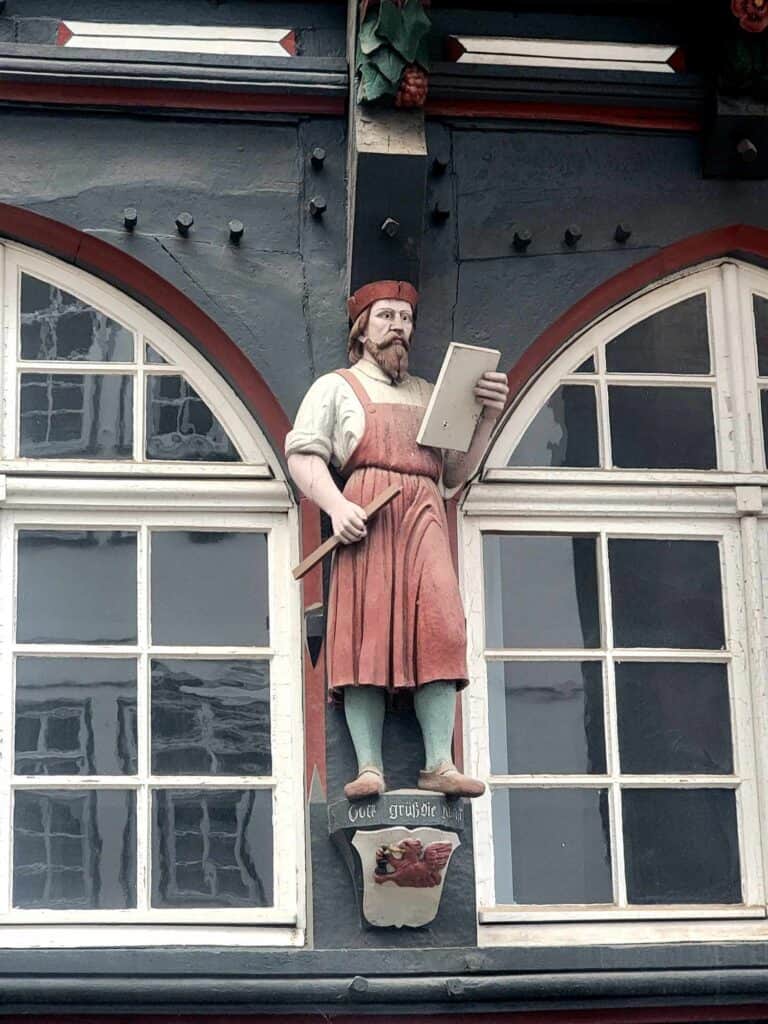 Carving of a man in a red hat and a matching apron holding a wooden stick and a book