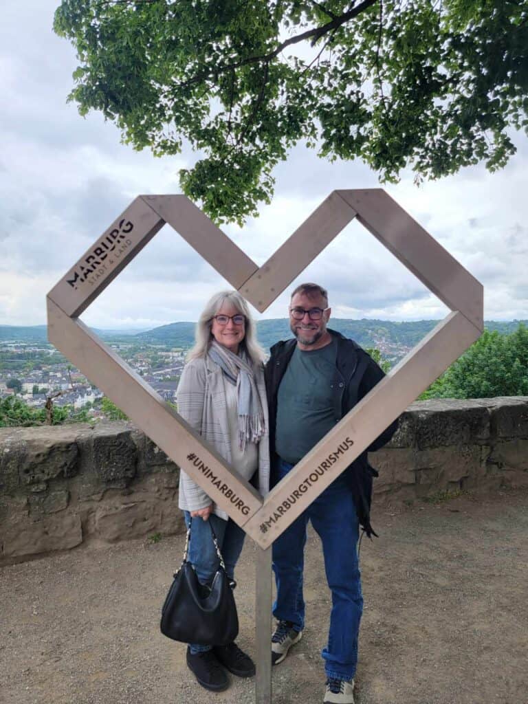 A couple stands on a terrace, framed in a wooden heart with panoramic view of the German countryside in the background