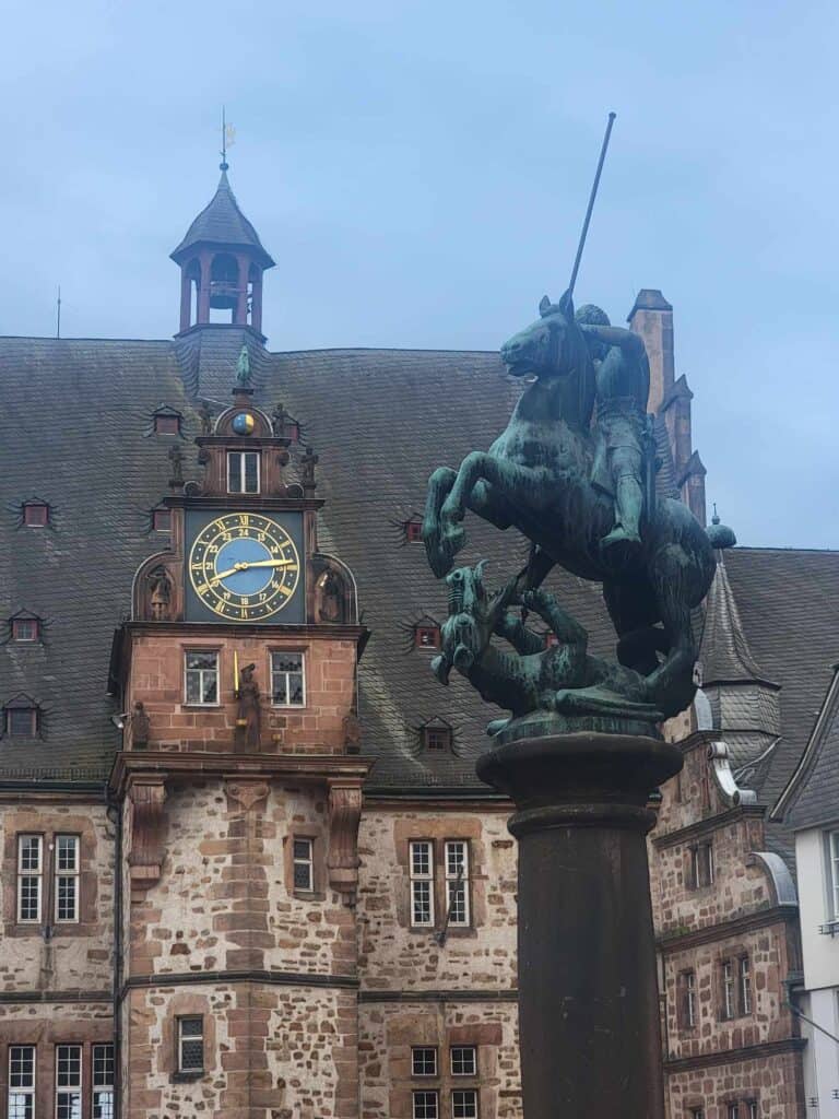 A stone building with a bell tower and blue clock. In front is a statue of St. George on horseback with a lance about to slay the dragon