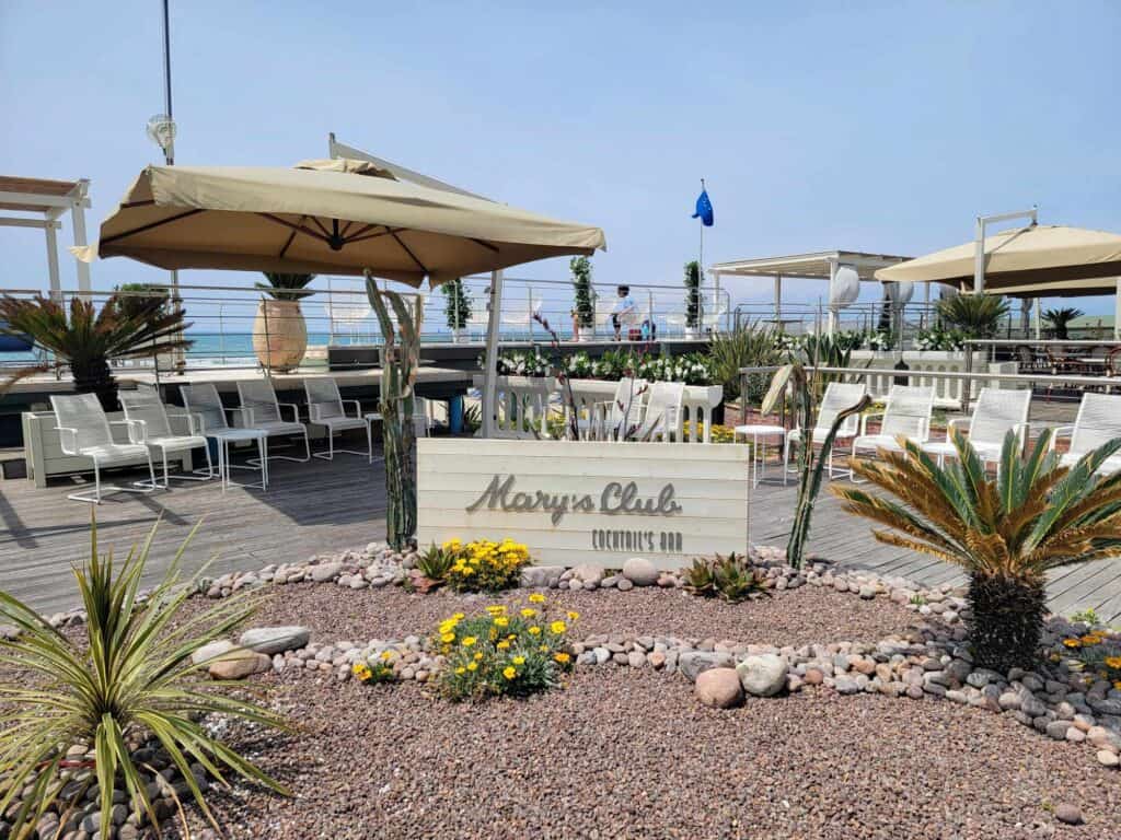 A sign that says Mary's Club & Cocktails outside with decorative plants, tables, chairs and umbrellas