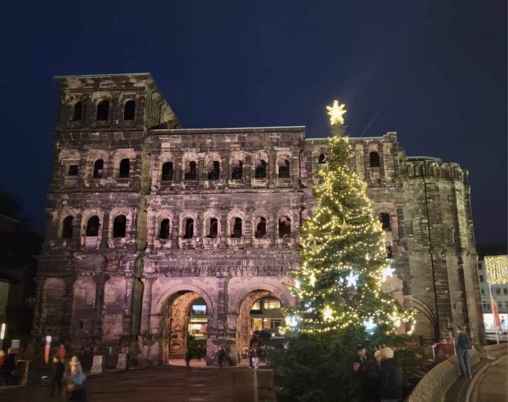 The large imposing Roman Porta Nigra with multiple stories of arches, made of dark stone, and an illuminated Christmas tree on the right