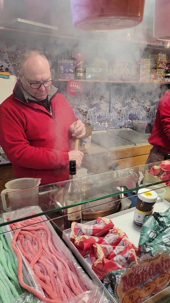 A man mixes a steaming bowl of candied nuts with paper cones full of nuts to sell in a glass case