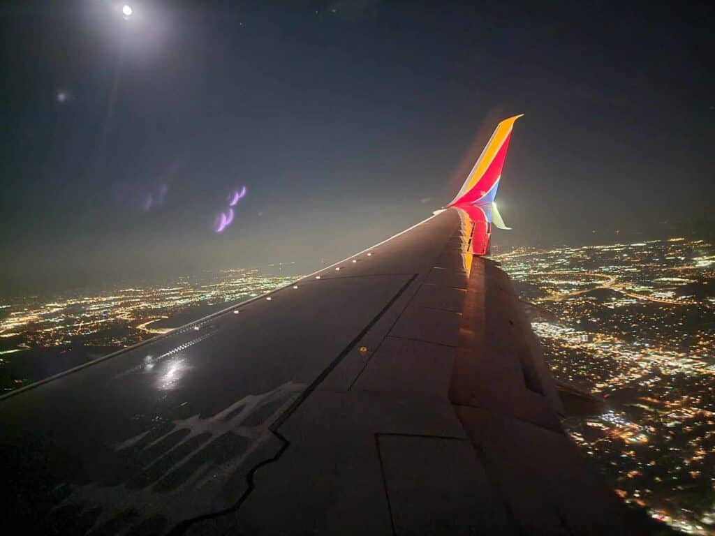 The wing of a Southwest aircraft with a red and orange tip flying over a city at night