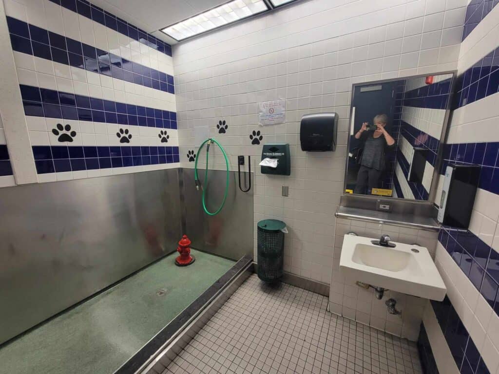 A pet relief area showing a fire hydrant, area for dogs to go, a waste basket, a poop bag dispenser, sink and paper towels. Pawprints cross the tiled wall