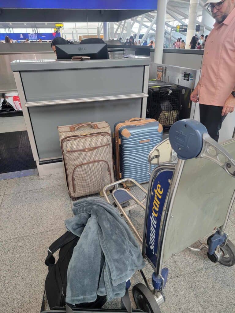 A cart and several suitcases. A large dog kennel sits on the scale at the check-in counter